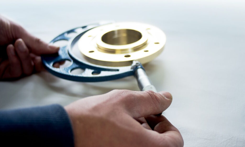 Close-up image of a technician using a micrometer caliper to measure the thickness of a precision-machined brass flange with multiple bolt holes