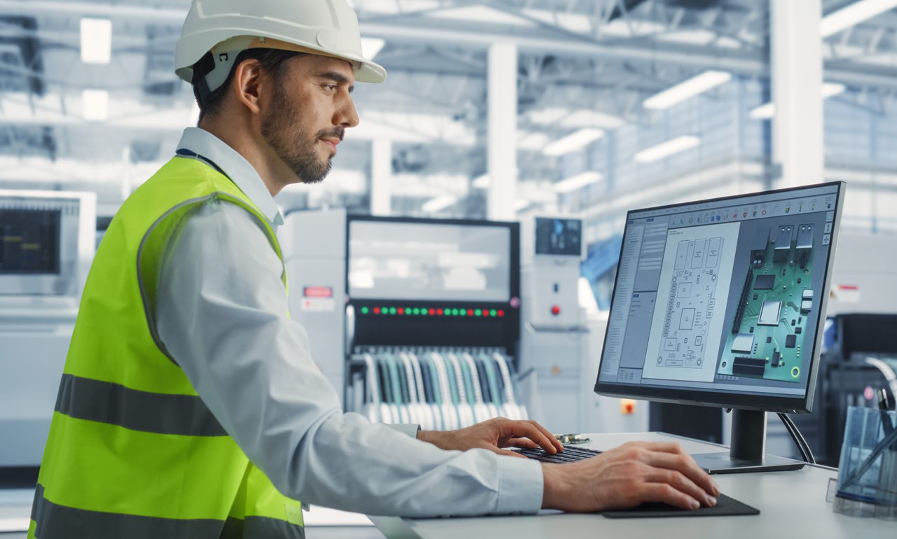 Engineer wearing a white safety helmet and high-visibility vest working on a desktop computer in a high-tech manufacturing facility, analyzing a 3D CAD model and circuit board layout design displayed on the screen