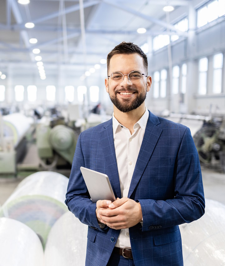 A smiling businessman with glasses and a tablet in a manufacturing facility